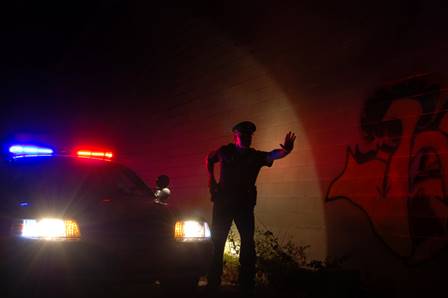 A silhouetted police officer holds up a hand in front of graffiti art near his police car with flashing lights. This image illustrates the article on wrongful shootings and civil rights violations.